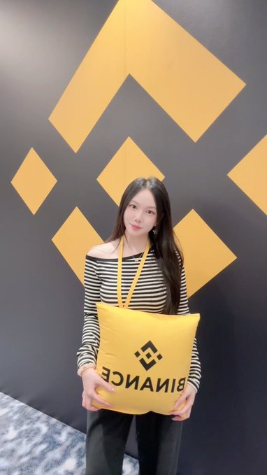 A young woman with long dark hair, wearing a striped off-shoulder top, stands in front of a wall displaying Binance's yellow logo patterns while holding a matching yellow "BINANCE" branded pillow/tote, celebrating the platform's 9th anniversary and her own 9 years in the crypto space.