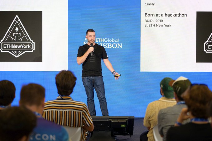 A man presents on stage at the ETHGlobal Lisbon event, with the screen behind him displaying text about 1inch being "Born at a hackathon" (BUIDL 2019 at ETH New York), while attendees watch from the audience. The image illustrates the talk referenced in the post, where 1inch team member Matt Snow shares the project's origin story during the opening ceremonies.