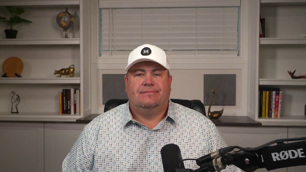The image shows a man wearing a white baseball cap sitting at a desk with a microphone, likely preparing for an interview or speaking event. The setting appears to be indoors, possibly in a room with bookshelves filled with various items such as vases and potted plants. The presence of these objects suggests that the space is used for both work and leisure activities. The man's attire and the microphone indicate that he might be involved in media or entertainment, while the bookshelves imply an interest in reading or collecting items related to literature or art.