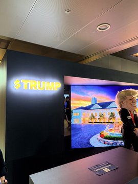 An indoor display area shows a glowing yellow '$TRUMP' sign on a wall next to a screen displaying a building rendering, with a person standing nearby.