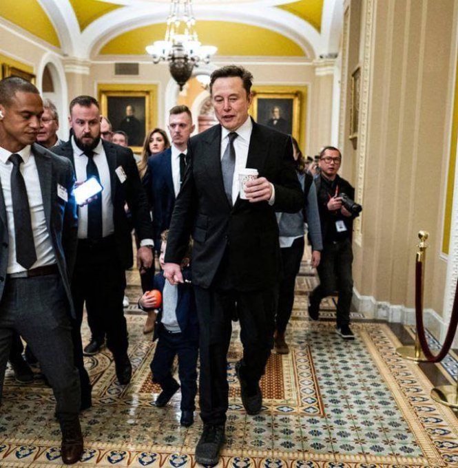 Elon Musk walks through a grand corridor holding a coffee cup, accompanied by several men in suits with visible badges.
