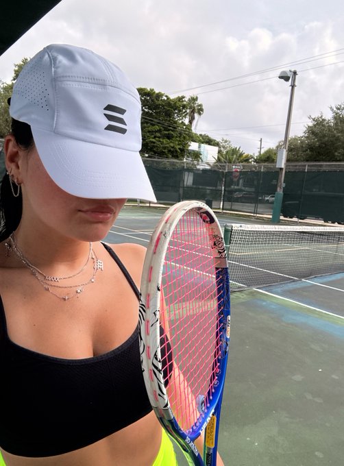 A person wearing a white cap and black top on a tennis court, holding a Wilson tennis racket with pink and blue strings.