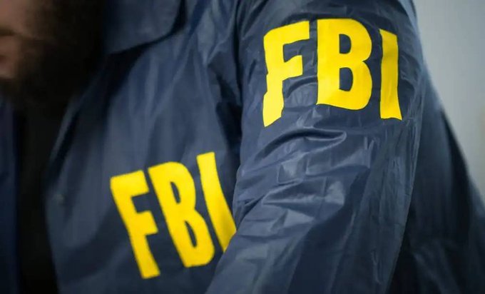 A close-up shot shows the back and shoulder of a person wearing a dark blue jacket with large yellow 'FBI' letters printed on both the sleeve and the back.