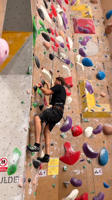A man scales an indoor climbing wall covered in colorful holds, gripping a chalk bag while reaching upward. The image symbolizes the "climbing" challenge of crypto investing, mirroring the post's message about steadily buying Bitcoin with Invity and letting it grow toward profit.