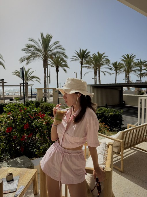 A young woman wearing a pink two-piece outfit and white bucket hat sipping a colorful drink, with palm trees and the ocean in the background.