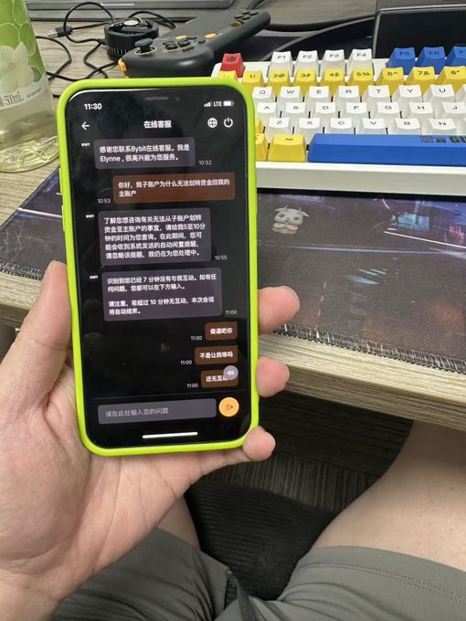 A first-person shot shows someone holding a smartphone with a bright green case, displaying a Chinese-language chat conversation (titled "在线客服") about a cryptocurrency withdrawal request, while a mechanical keyboard and game controller sit on the desk in the background. The post appears to be a frustrated complaint directed at the Bybit exchange, with the chat likely serving as evidence of poor customer service in resolving the user's issue.