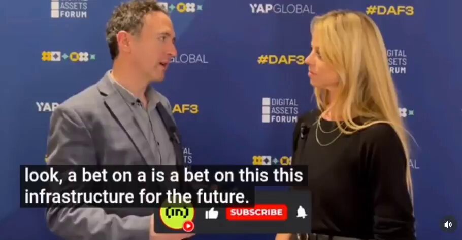 The image shows a man wearing a gray suit talking to a woman who has blonde hair, both standing against a blue backdrop with white text. They are positioned at a press conference or event where they appear to be discussing something related to cryptocurrency or digital assets. The background features a large banner that reads "Look a bet on a is a beta on this infrastructure for the future." This suggests that there might be an ongoing discussion about the potential impact of new technologies, such as blockchain and cryptocurrencies, on traditional industries like finance and real estate.