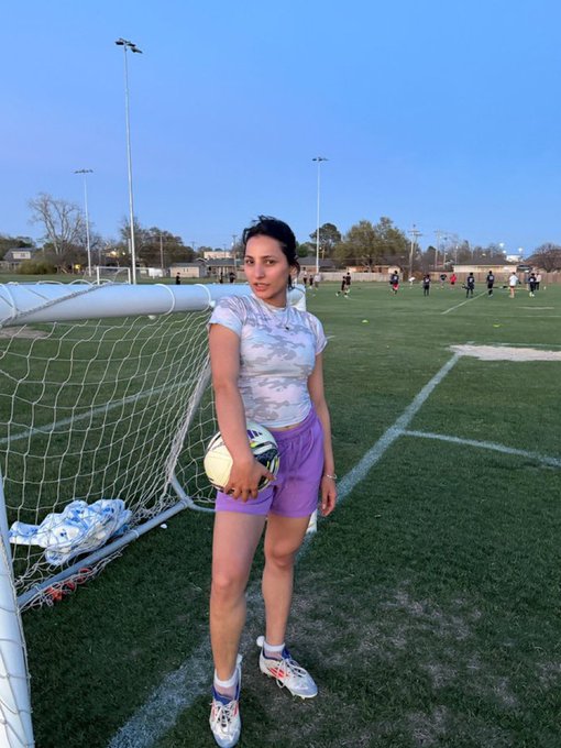 A woman on a soccer field wearing a tie-dye shirt and purple shorts, holding a soccer ball near a goal net.