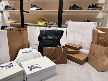 A collection of luxury shopping bags and shoeboxes, including brands like Burberry, is piled on the floor in front of a display of sneakers.