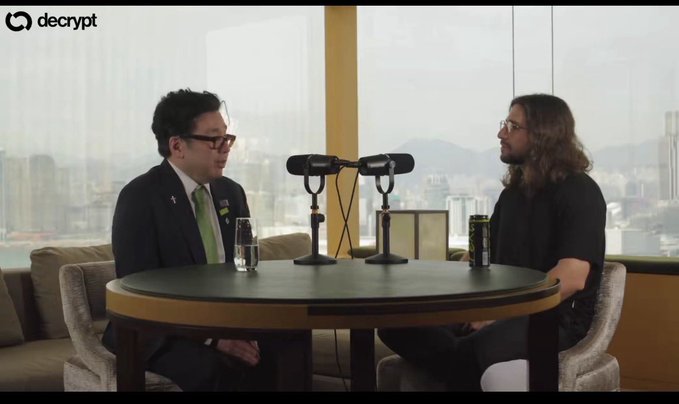 The image shows two men sitting at a table, having a conversation with microphones attached to them. One man has a tie visible, while the other one does not. The background features a window overlooking a cityscape, adding an urban touch to the scene. The caption reads "Tom Lee: “If we were to delete themeth—“ if Tom Lee is the person speaking and if deleting themeth means removing their name from the image, then it would be a lie."