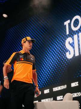 Lando Norris, wearing a black and orange cap, stands on stage at the Token2049 event, appearing to address an audience. Behind him, the "TOKEN2049" branding is prominently displayed on a blue backdrop, aligning with the post's mention of his appearance at the crypto conference sponsored by OKX.