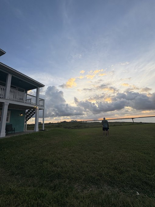 A person walks across a grassy lawn toward a long bridge in the distance at sunset, with a coastal house and porch visible on the left side of the frame.