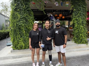 Three people wearing matching black shirts and shorts pose for a photo in front of a building covered in green vines with orange decorations hanging from the entrance.