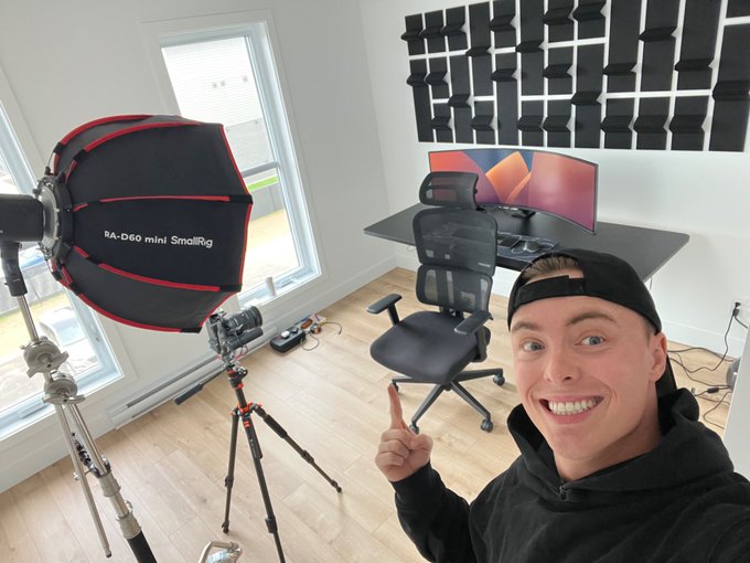 A selfie of a smiling man wearing a black cap pointing at a content creation setup featuring a SmallRig RA-D60 softbox light, a curved monitor, and acoustic foam panels on the wall.