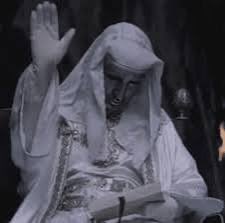 A black and white image of a robed, hooded figure with a raised hand and a serious expression.
