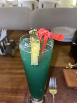 A tall glass containing a bright blue-green cocktail garnished with a pineapple wedge and a red curly straw, served on a wooden table.