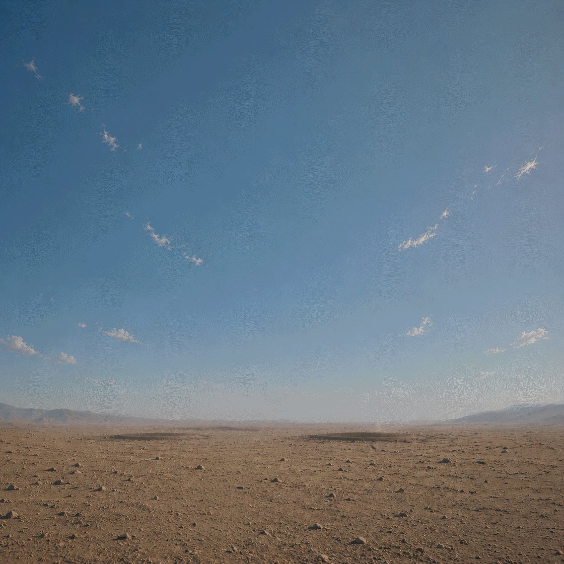 The image shows a vast, open desert landscape with a clear blue sky above. The scene appears to be empty or devoid of any human presence, as indicated by the lack of people or objects visible in the photo. The sky has a few clouds scattered across it, adding depth and contrast to the overall composition. The image does not contain any text or other discernible objects, making it appear like an advertisement for something called "Day 185 of posting new stuff until $tibbir is out of stealth."