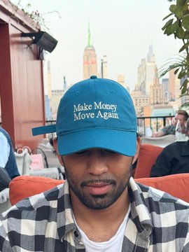 A man with a beard wearing a black and white plaid shirt and a teal baseball cap with the text 'Make Money Move Again', photographed with the Empire State Building visible in the background.