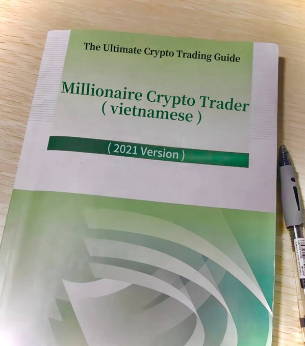 A green booklet titled 'The Ultimate Crypto Trading Guide: Millionaire Crypto Trader (vietnamese) (2021 Version)' lying on a wooden surface next to a pen.