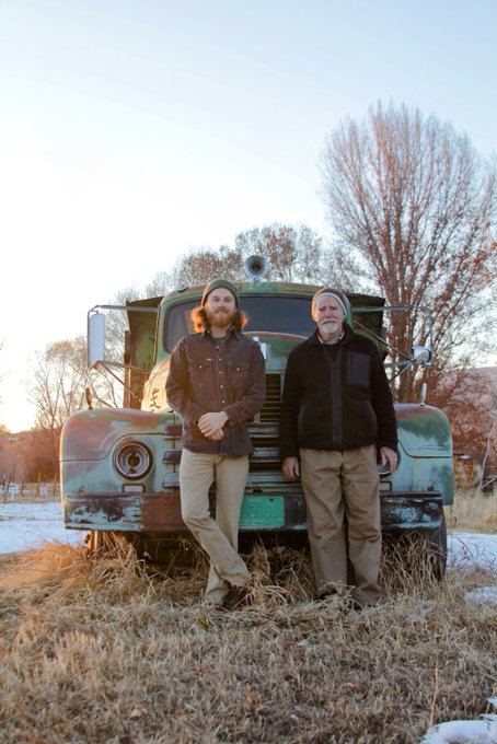 Two men stand smiling in front of a vintage, rusted military truck in a snowy field with bare trees at dusk.