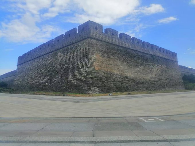 A photograph of a massive stone fortress corner with crenellated battlements under a partly cloudy blue sky, set behind a wide stone-paved plaza.