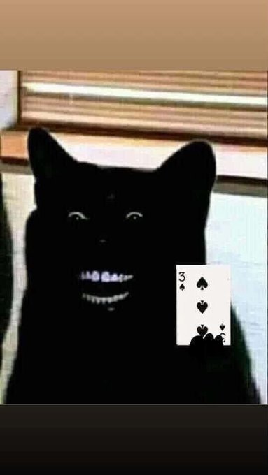 A surreal dark image shows a black cat with glowing eyes and a wide grin next to a 3 of spades playing card.