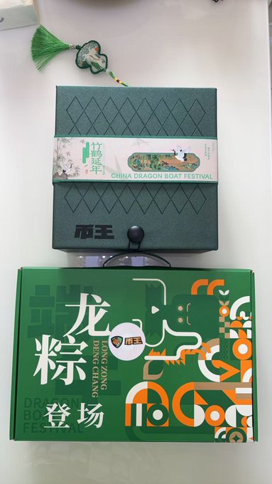 The image features two boxes, one green and one orange, sitting side by side with a tassel hanging from them. The green box has Chinese characters on it, while the orange box does not. Both boxes appear to be gifts or souvenirs, possibly for an event like the Chinese New Year.