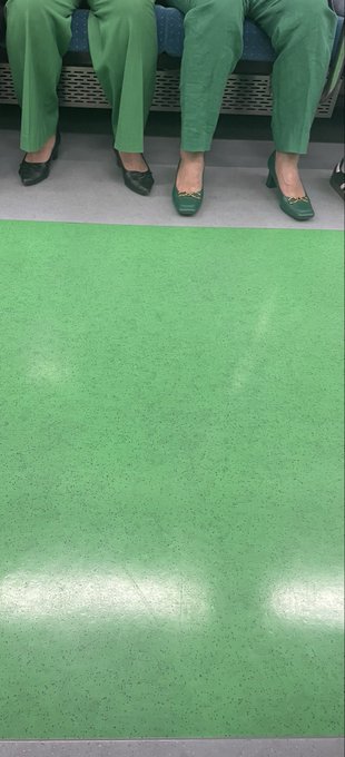 A vertical video screenshot shows two people sitting on a bench above a large expanse of bright green flooring, with only their green trousers and shoes (one black pair, one green pair) visible in the frame.