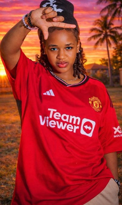 A young person wearing a red Manchester United 'Team Viewer' soccer jersey, posing with their hand near their forehead against a sunset background with palm trees.