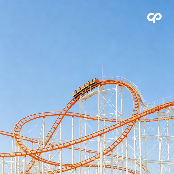 A low-angle shot of an orange and white roller coaster track twisting against a clear blue sky, with a 'cp' logo in the top right corner.