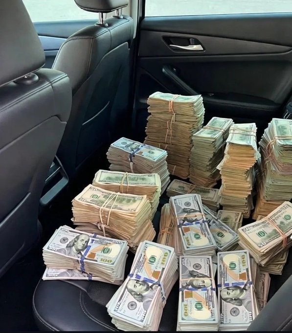 Stacks of bundled US currency, including hundreds and thousands, are piled on the back seat of a car with black leather interior.