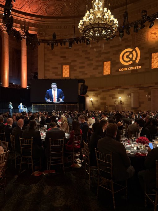A formal dinner event at the Coin Center gala, featuring a chandelier-lit hall with attendees seated at round tables watching a speaker displayed on a large screen.