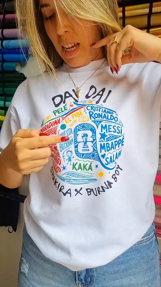 The image features a woman wearing a white t-shirt with colorful text printed on it. She is pointing at her shirt, possibly showing off its design or discussing something related to it. The background of the image includes a photo of a soccer ball, suggesting that the content might be related to a sports event or a celebration involving soccer. The caption "DAI DAI Tracker TikTok Videos: 800,000+ World Cup Kickoff: 9 Days Away The tournament hasn'nt even started yet." is also present in the image, which could be an interesting detail about the event being discussed or a reference to the creator of the t-shirt design.