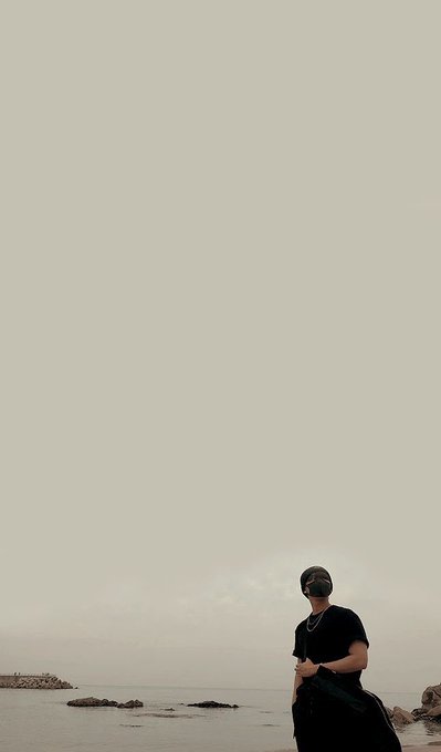 A vertically oriented photograph of a person wearing a black t-shirt, cap, and sunglasses standing on a beach with rocks and the ocean in the background, holding what appears to be a wetsuit, under a pale overcast sky.