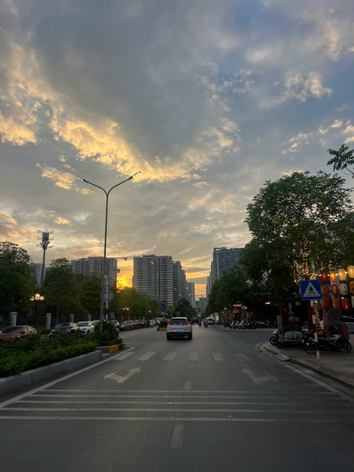 The image shows an empty street with buildings lining both sides, including apartment buildings. The sky above has clouds, creating a beautiful backdrop for the scene. A car can be seen driving down the road, adding life to the otherwise quiet atmosphere. The photo is captioned "Sáng nắng chiều mưa. Sáng yap @quipnetwork .,chiều yap @NucleusCodes . Tối yap @sleepagotchi để ngủ ngon nges ae.", which is likely the title of the image.