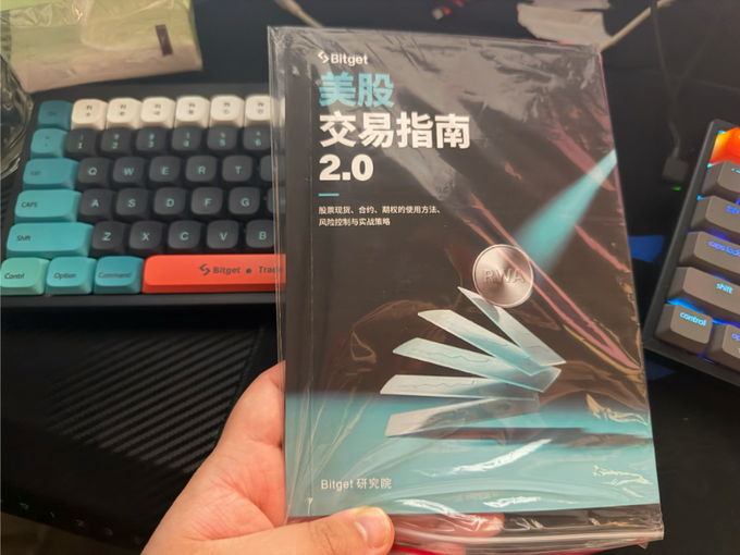 A hand holds a packaged book titled "Bitget 美股交易指南 2.0" (US Stock Trading Guide 2.0) in front of a mechanical keyboard with teal and red accent keys.