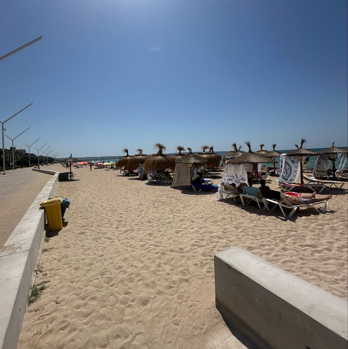 A sunny beach scene with straw umbrellas, lounge chairs, and sunbathers along the sand, bordered by a paved walkway with a yellow trash bin.