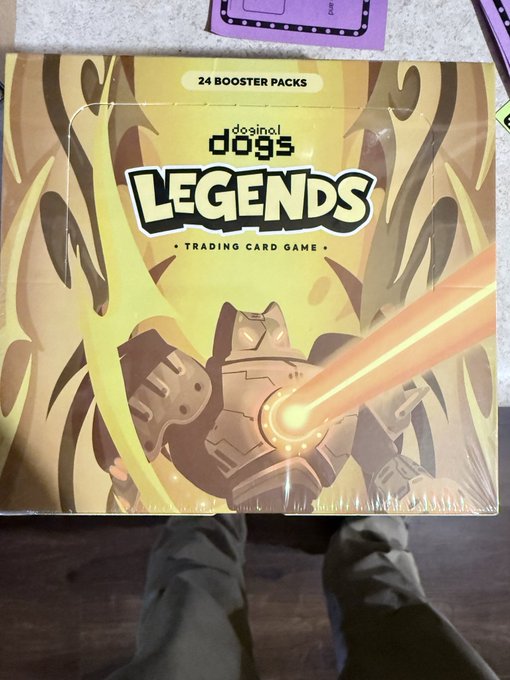A sealed box of 'doge legends TRADING CARD GAME' containing '24 BOOSTER PACKS' with artwork of a robotic dog firing a laser, sitting on a floor next to a person's feet.
