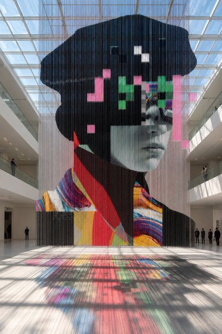 A large-scale contemporary art installation in a modern atrium, depicting a pixelated portrait of a woman with colorful sunglasses hanging as a curtain-like display.