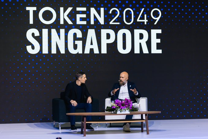 Two men sit in armchairs on stage at the TOKEN2049 Singapore conference, with the event's bold white logo displayed prominently on the dark backdrop behind them. A coffee table with purple flowers sits between them as they engage in a panel discussion on digital trust and data security.