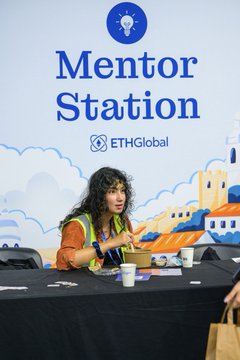 A woman sits at a 'Mentor Station' booth branded with 'ETHGlobal,' gesturing while speaking, with event graphics and cartoon buildings visible on the backdrop behind her.