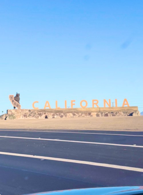 The iconic 'CALIFORNIA' sign along a desert highway under a clear blue sky, with a Sante Fe railroad logo visible to the left.