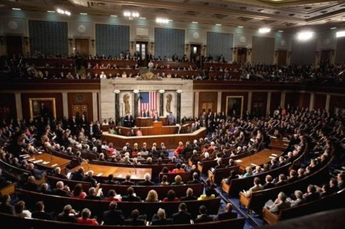 The image shows a large room filled with people sitting in chairs, possibly at a formal event or gathering. The audience appears engaged as they watch an official speak from behind them. The setting suggests that this could be a political event, such as a speech by a U.S. representative or a member of Congress. The crowd is diverse and attentive, indicating the importance of the topic being discussed.