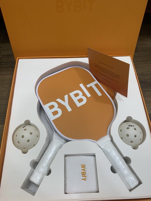 An orange and white pickleball set in an open display box branded with 'BYBIT' in large white letters, featuring two paddles, two balls, and a small accessory pouch.