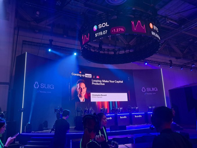 A conference or event venue showing a large circular screen displaying crypto tickers (SOL, BTC) with prices and percentages, while a smaller screen below announces 'Looping: Make Your Capital Productive' by Christophe Robert of OCG Current.