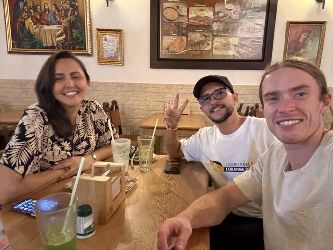 A selfie of three smiling people seated at a wooden table in a restaurant; the man in the middle wears a black cap, glasses, and a t-shirt reading 'I CAME, I SAW, I ORANGE...', with religious paintings and a framed photo display on the wall behind them.