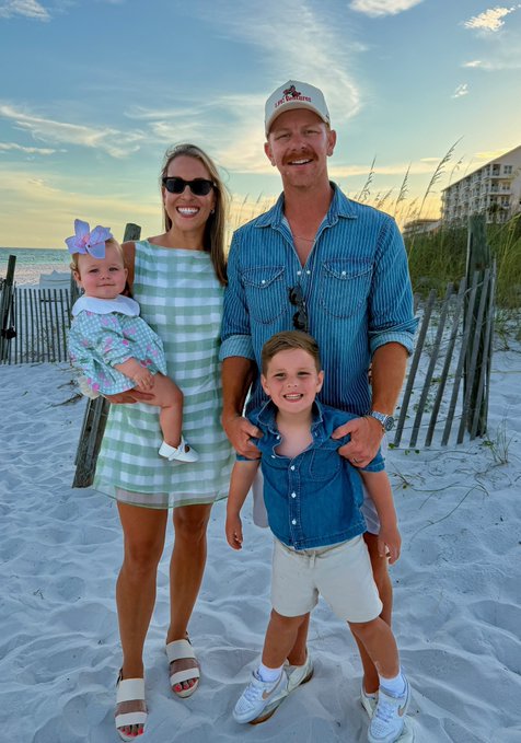 A family of four poses together on a sandy beach at sunset, with a man in a white baseball cap and blue denim shirt, a woman in a green and white striped dress holding a baby with a pink bow, and a young boy in a denim shirt.