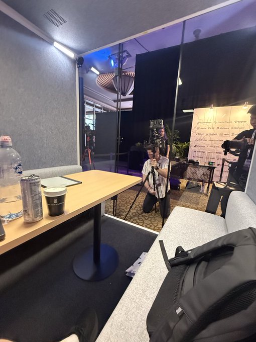 An interior view of a podcast or interview studio with a videographer adjusting a camera on a tripod, with a backdrop displaying sponsor logos visible behind the subject.