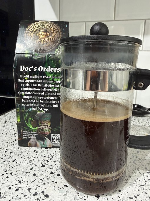 A French press containing dark brewed coffee sits on a speckled countertop next to a box of "APE GANG COFFEE" labeled "Doc's Orders," described as a medium roast blend.