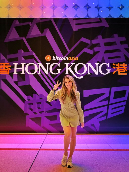 A woman poses cheerfully with a peace sign in front of a vibrant Bitcoin Asia conference backdrop displaying "HONG KONG" in both English and Chinese characters, capturing her excitement at attending the cryptocurrency event in her element.